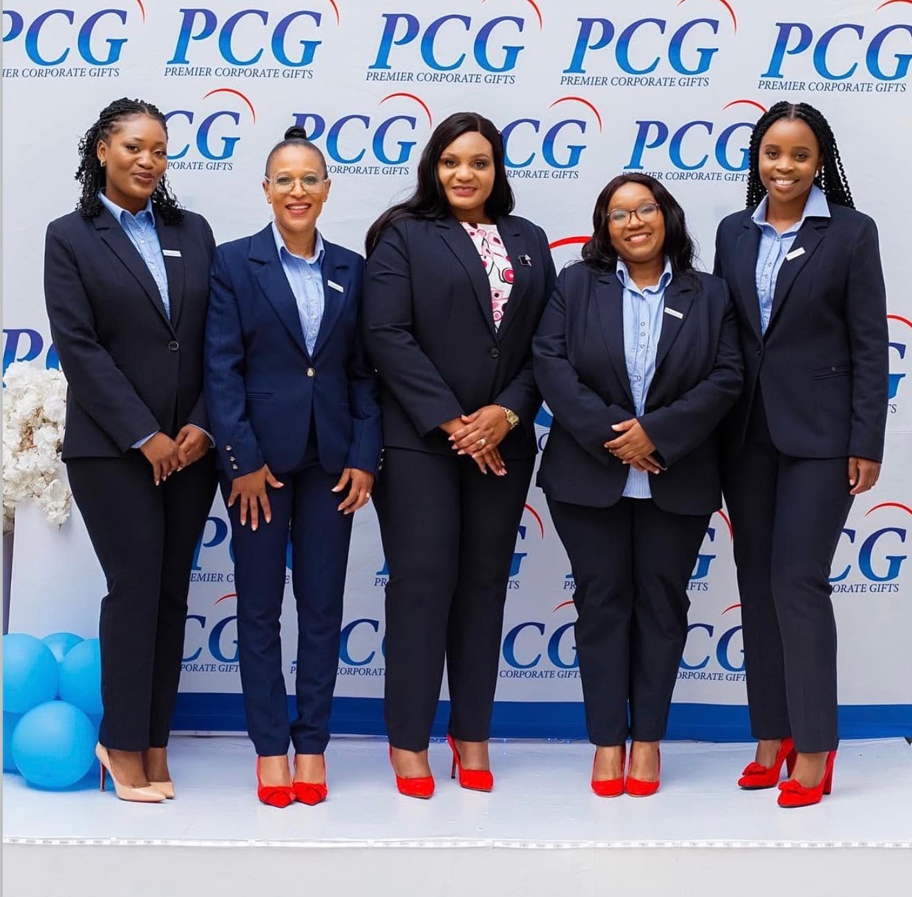 Five Premier Corporate Gifts team members in matching navy suits smiling in front of a PCG step-and-repeat backdrop