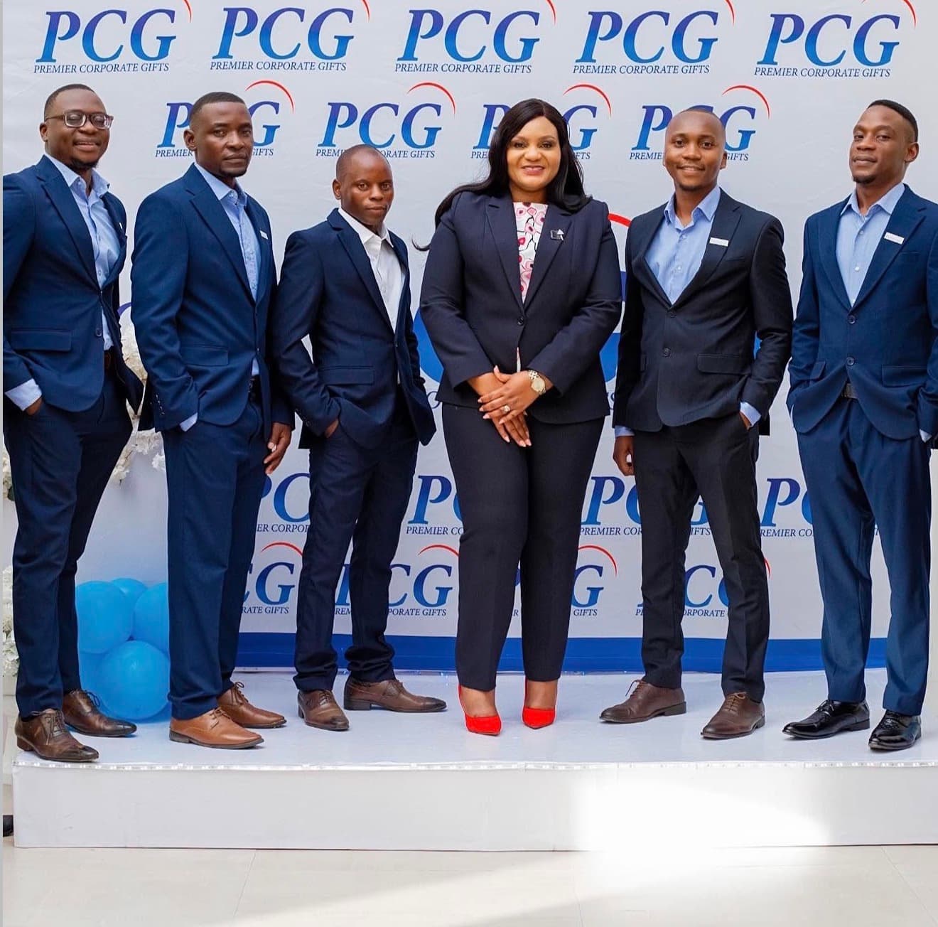 Premier Corporate Gifts colleagues in navy suits standing together on a white platform before a branded backdrop
