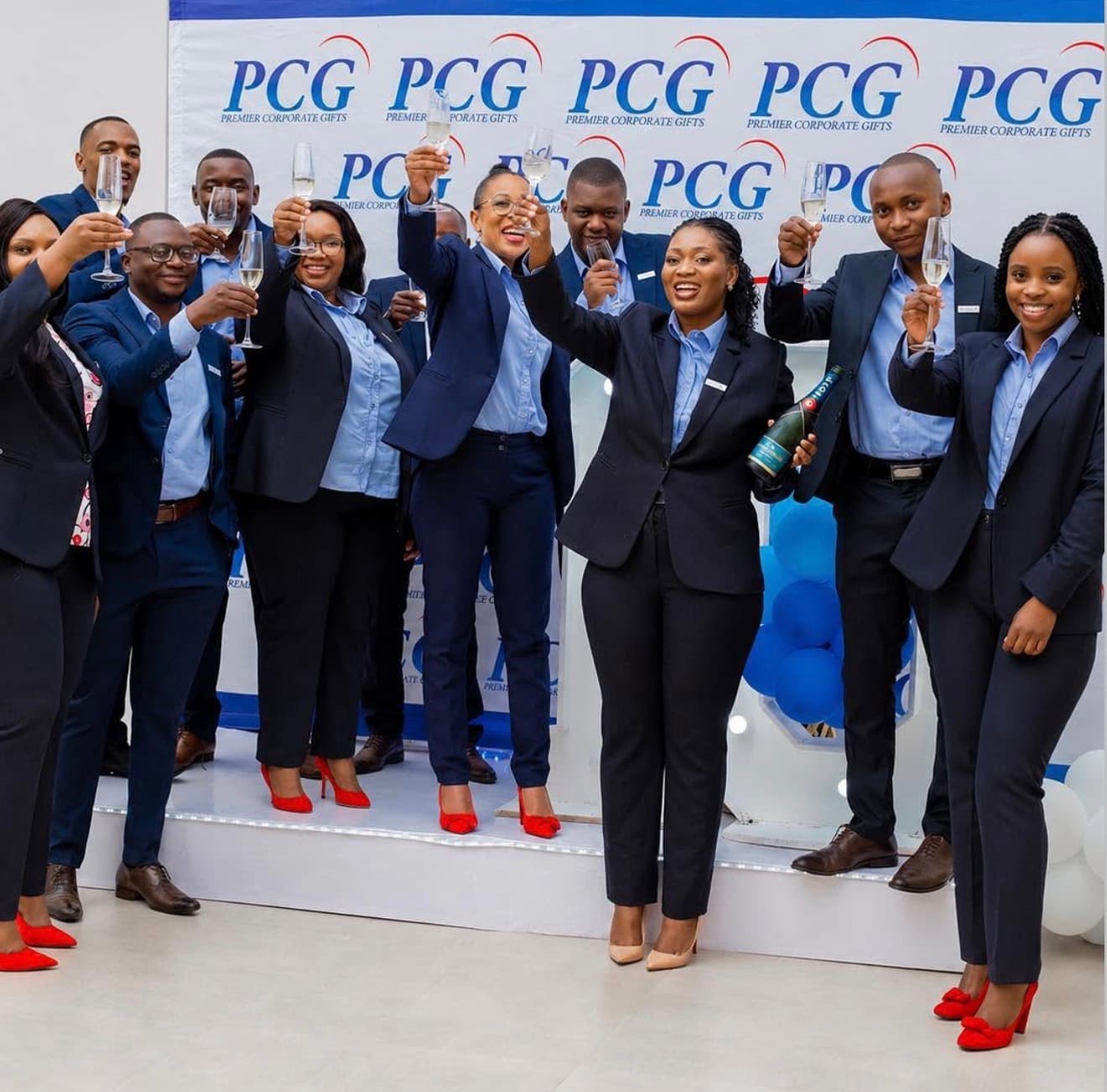 Premier Corporate Gifts team raising champagne glasses in a celebratory toast before a branded PCG backdrop