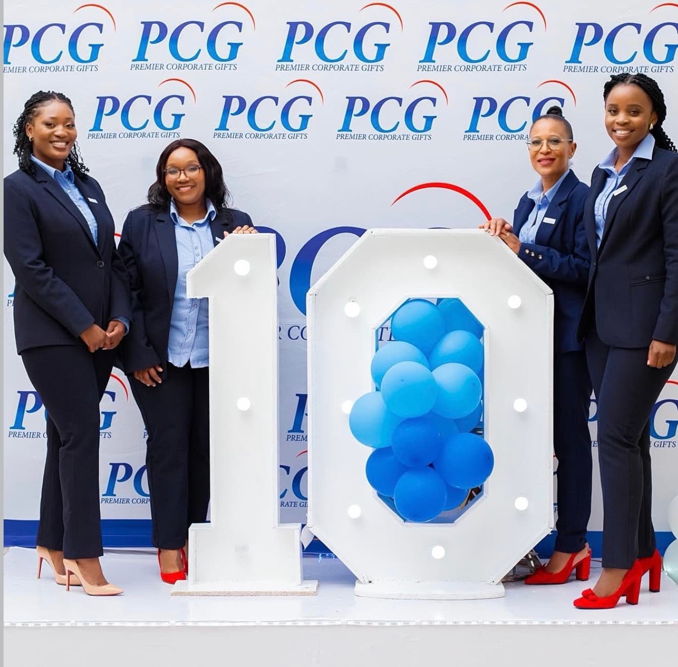 Four Premier Corporate Gifts team members standing beside a large white number display with blue balloons
